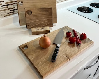 May include: Three rectangular wooden cutting boards with circular corner holes, one featuring a small wooden stand with the text DAAK. A stainless steel chef knife, a yellow onion, three red cherry tomatoes, and a red chili pepper are present.