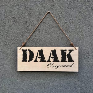 Könnte beinhalten: Ein rechteckiges Holzschild mit dem Wort "DAAK" in fetten schwarzen Buchstaben und dem Wort "Original" in Kursivschrift. Das Schild ist an einem braunen Seil an einer strukturierten grauen Wand aufgehängt.