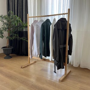 May include: A wooden clothing rack with several garments hanging on it. The garments include a striped shirt, a white shirt, a green shirt, a cream-colored shirt, and a dark gray trench coat. The rack is made of light-colored wood and has a simple, minimalist design.