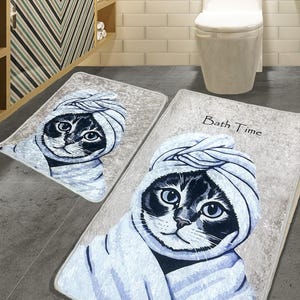 K&ouml;nnte beinhalten: Zwei Badematten mit einem Katzendesign. Die gr&ouml;&szlig;ere Matte hat den Text "Bath Time" darauf. Die Katze tr&auml;gt ein Handtuch und hat einen s&uuml;&szlig;en Gesichtsausdruck.