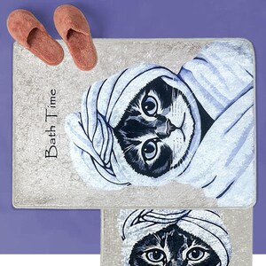 K&ouml;nnte beinhalten: Zwei Badematten mit grauem Hintergrund und einer Schwarzwei&szlig;illustration einer Katze, die ein Handtuch tr&auml;gt. Die obere Matte hat den Text "Bath Time" aufgedruckt.