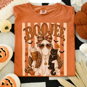 May include: A burnt orange t-shirt featuring a graphic of a ghost wearing a plaid bow, heart-shaped sunglasses, and layered necklaces. The ghost holds a pumpkin-themed iced coffee and stands next to a black cat. The text reads BOOJEE.