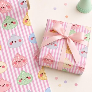 May include: Wrapping paper roll and a gift box wrapped in paper featuring a pattern of cute, smiling, glittery dumpling characters on a background of vertical pink and white stripes. The gift box is tied with a light pink satin ribbon bow.