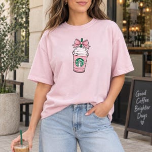 Könnte beinhalten: Ein hellrosa T-Shirt mit kurzen Ärmeln und einer Grafik eines rosa Kaffeebechers mit Schlagsahne, einem grünen Strohhalm und einer rosa Schleife. Der Becher zeigt ein kreisförmiges grün-weißes Logo mit einer Sirenenfigur in der Mitte.