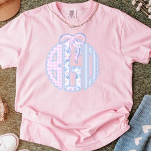 May include: A light pink short-sleeve t-shirt featuring a circular monogram graphic in the center. The monogram consists of three letters with distinct patterns: a pink gingham check, a blue floral print, and blue vertical stripes, topped with a light pink bow.