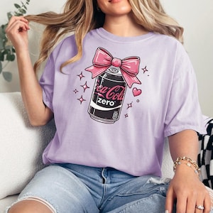 May include: A lilac t-shirt with a graphic of a Coca-Cola Zero can adorned with a pink bow, hearts, and stars. The Coca-Cola Zero can is black and white with pink branding.
