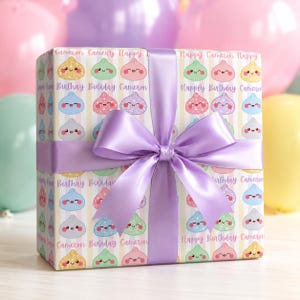 May include: A rectangular gift box wrapped in paper featuring a repeating pattern of smiling, glittery cartoon figs in pastel colors, interspersed with the text Happy Birthday Cameron, tied with a wide, smooth, lavender-colored satin ribbon bow.