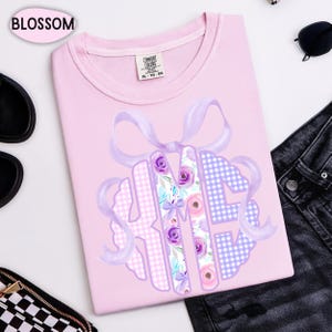 May include: A light pink t-shirt with a large monogram design. The monogram features the letters H, K, and M, decorated with floral and gingham patterns in shades of purple, pink, and blue. A light purple ribbon bow is at the top of the monogram.