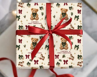 May include: A square gift box wrapped in cream-colored paper featuring a repeating pattern of teddy bears holding gifts, rocking horses, red bows, and green presents. The text 'CARTER'S FIRST CHRISTMAS' is printed below each bear. A red satin ribbon is tied in a bow.