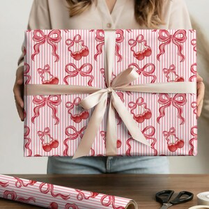 以下が含まれることがあります： ピンクと白のストライプの包装紙で包まれたギフト。赤いリボンとさくらんぼの模様があります。ギフトはベージュのリボンとリボンで結ばれています。テーマはギフト包装です。
