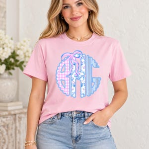Puede incluir: Una camiseta de manga corta de color rosa claro con un gran gráfico de monograma circular en el pecho. El monograma consta de tres letras en tonos azul claro y rosa, incorporando patrones de cuadros vichy, flores y rayas con un lazo.