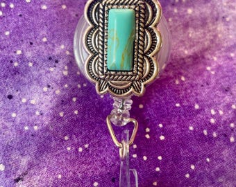 May include: A retractable badge reel featuring a rectangular turquoise stone with yellow veining, set in a decorative silver-toned metal frame with a scalloped edge and rope-like detailing, attached to a clear plastic strap with a metal snap button.