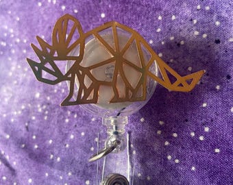 May include: A gold-colored, geometric triceratops dinosaur badge reel. The dinosaur is attached to a clear plastic reel with a metal clip and a retractable cord. The background is a purple fabric with white speckles.