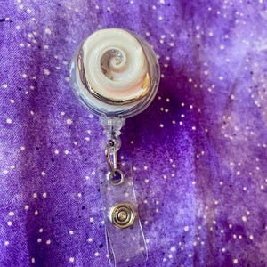 May include: A clear, retractable badge reel with a white and silver spiral design inside. The reel has a clear plastic strap with a metal snap closure. The background is a purple fabric with white polka dots.