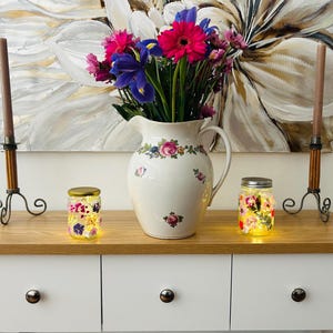 Può includere: Due barattoli di vetro con disegni floreali e luci all'interno, posizionati su un tavolo di legno con una cassettiera bianca dietro. Un'anfora bianca con disegni floreali si trova tra i barattoli. Un mazzo di fiori rosa, viola e bianchi è posto nell'anfora. Due candelabri marroni con basi in metallo nero si trovano su entrambi i lati dei barattoli.