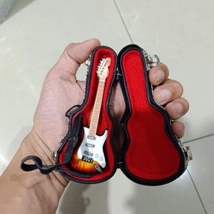 Può includere: Miniatura di chitarra elettrica bianca e gialla in una custodia nera aperta con interno rosso. La chitarra ha un corpo bianco, manico marrone e hardware argentato. La custodia è aperta, rivelando la chitarra.