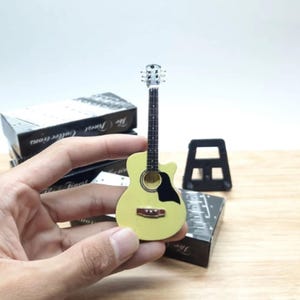 Puede incluir: Una peque&ntilde;a guitarra ac&uacute;stica de color amarillo claro con un golpeador negro y una boca marr&oacute;n. La guitarra se sostiene en una mano, con un soporte negro y una caja en el fondo. La guitarra es un objeto de colecci&oacute;n.