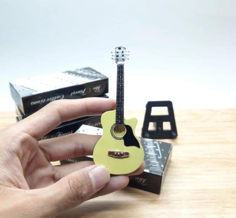Puede incluir: Una peque&ntilde;a guitarra ac&uacute;stica de color amarillo claro con un golpeador negro y una boca marr&oacute;n. La guitarra se sostiene en una mano, con un soporte negro y una caja en el fondo. La guitarra es un objeto de colecci&oacute;n.