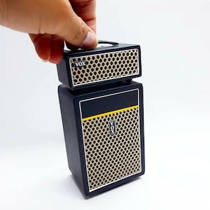Op de afbeelding: Een miniatuur zwarte en gouden VOX-versterkerstapel. De versterker heeft een zwarte textuurafwerking met gouden mesh luidsprekerhoezen. De bovenste versterker heeft een handvat. Het VOX-logo is zichtbaar op beide versterkers.