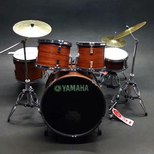 Puede incluir: Batería en miniatura con un bombo negro con el logotipo de Yamaha en verde. El juego incluye tambores de madera, platillos y soportes cromados. Los platillos son de color dorado. La batería es una réplica a pequeña escala.