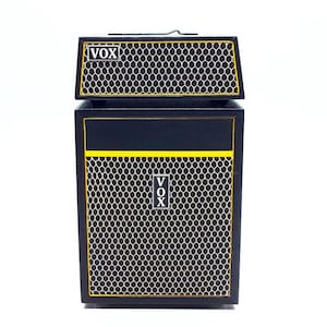 Könnte beinhalten: Ein schwarzer und gelber Miniaturverstärker mit dem Wort "VOX" auf der Vorder- und Oberseite. Der Verstärker hat ein wabenförmiges Grilldesign und ist wahrscheinlich ein Dekorationsartikel oder ein Spielzeug.