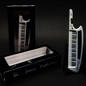 May include: A silver and black AX-Synth keyboard with a black and white keyboard design. The keyboard is displayed next to its black box, which reads "The Finest Collections". The keyboard is a musical instrument.