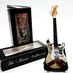 May include: A miniature electric guitar with a white body and colorful floral design. The guitar is on a black stand and has a black box with the text "The Finest Collection".