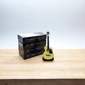 Puede incluir: Una peque&ntilde;a guitarra ac&uacute;stica amarilla con detalles negros se exhibe sobre un soporte negro. Junto a ella, cuatro cajas negras con texto blanco, incluyendo "The Finest Collections" y "The Mini Guitar World".