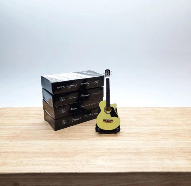 Puede incluir: Una peque&ntilde;a guitarra ac&uacute;stica amarilla con detalles negros se exhibe sobre un soporte negro. Junto a ella, cuatro cajas negras con texto blanco, incluyendo "The Finest Collections" y "The Mini Guitar World".