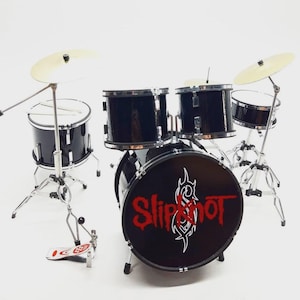 Może przedstawiać: Miniaturowy czarny zestaw perkusyjny ze srebrnym osprzętem i talerzami. Bęben basowy zawiera nazwę zespołu "Slipknot" w czerwonych i białych literach. Mała, szczegółowa replika zestawu perkusyjnego.