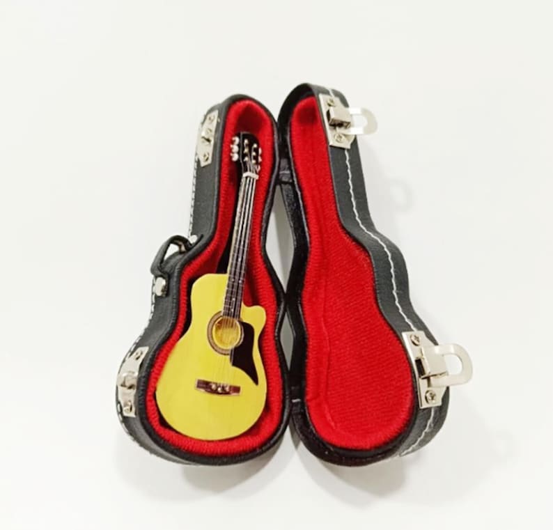 Puede incluir: Guitarra ac&uacute;stica en miniatura en un estuche negro con forro rojo. La guitarra tiene un cuerpo amarillo y cuerdas plateadas. El estuche est&aacute; abierto, mostrando los detalles de la guitarra. Un objeto de colecci&oacute;n encantador.