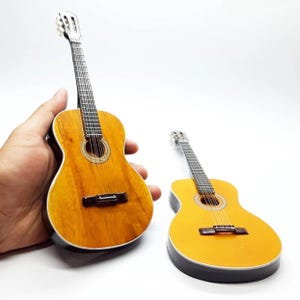 以下が含まれることがあります： 2つのミニチュアのアコースティックギター。1つは手で持たれており、天然の木目仕上げが施されています。もう1つのギターは明るい黄色です。どちらも黒いブリッジと指板があり、銀色のペグが付いています。