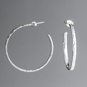 A pair of argentium sterling silver hammered post hoop earrings.  The earrings are small and lightweight, with a simple, minimalist design. The textured metal gives them a unique, handcrafted look. These stylish earrings are perfect for everyday wear or a special occasion. The hoops are slightly irregular in shape, reflecting the hammered texture.  They are shown against a neutral gray background.