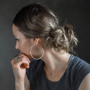 May include: A woman wearing large, textured silver hoop earrings. The earrings are circular and have a hammered or textured appearance. She is wearing a gray top and has her hair pulled back in a bun.