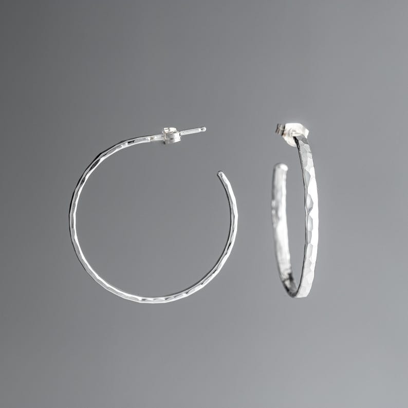 A pair of argentium sterling silver hammered post hoop earrings.  The earrings are small and lightweight, with a simple, minimalist design. The textured metal gives them a unique, handcrafted look. These stylish earrings are perfect for everyday wear or a special occasion. The hoops are slightly irregular in shape, reflecting the hammered texture.  They are shown against a neutral gray background.
