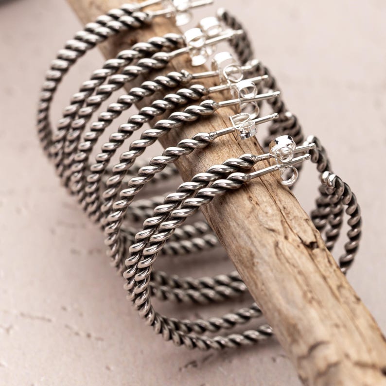 May include: A collection of silver-colored hoop earrings with a twisted rope design. The earrings are displayed on a wooden stick against a neutral background. The earrings have a post and clasp closure.