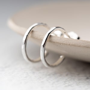 May include: A pair of small, silver hoop earrings. The earrings have a hammered texture and are displayed on a light-colored, wooden surface. The hoops are slightly curved and have a simple, minimalist design.