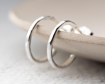 May include: A pair of small, silver hoop earrings. The earrings have a hammered texture and are displayed on a light-colored, wooden surface. The hoops are slightly curved and have a simple, minimalist design.