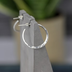 The side profile of these one inch sterling silver hoops shows the hand hammered texture and subtle flare that the hammer has given them.  They are perched on top of a gray concrete earring holder.  This view shows the post and ear nut.