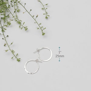 A pair of hammered, nickel free sterling silver hoop earrings are shown with an arrow for scale which indicates that they measure 1" across 25mm.  The earrings have a post ear wire and a secure butterfly ear nut to hold them in place.