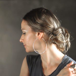 May include: A large, thin, circular silver hoop earring with a smooth, polished finish. The earring is worn in the earlobe, featuring a simple wire closure mechanism that connects the two ends of the circular metal band together.