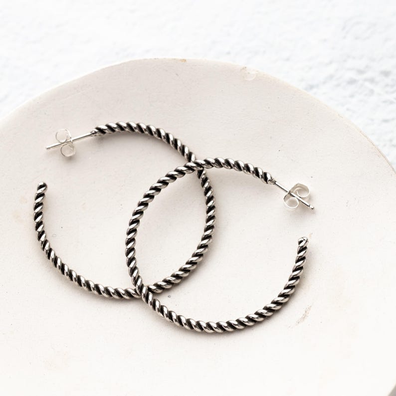 May include: A pair of silver hoop earrings with a twisted rope design. The earrings are displayed on a white, slightly textured surface. The earrings have a post and butterfly clasp.