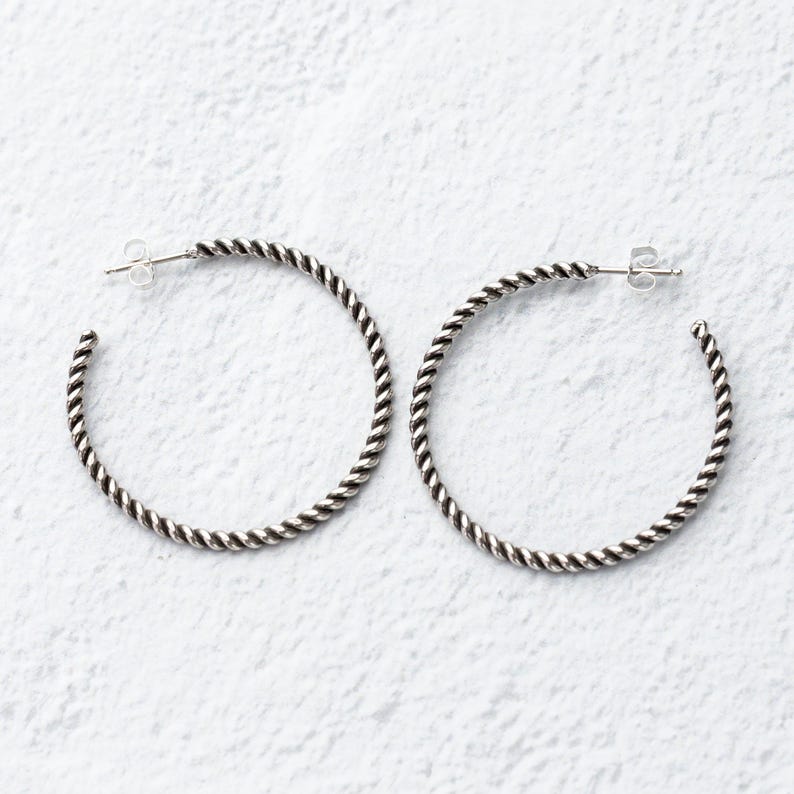 May include: A pair of silver-toned hoop earrings. Each earring has a twisted rope design around the hoop. The earrings are displayed on a white textured surface. The earrings have a post and butterfly clasp.