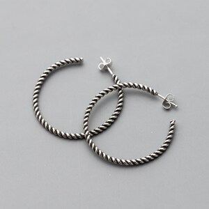 May include: A pair of silver hoop earrings with a twisted rope design. The earrings have a post and butterfly clasp closure. The hoops are a medium size and are displayed on a gray background.