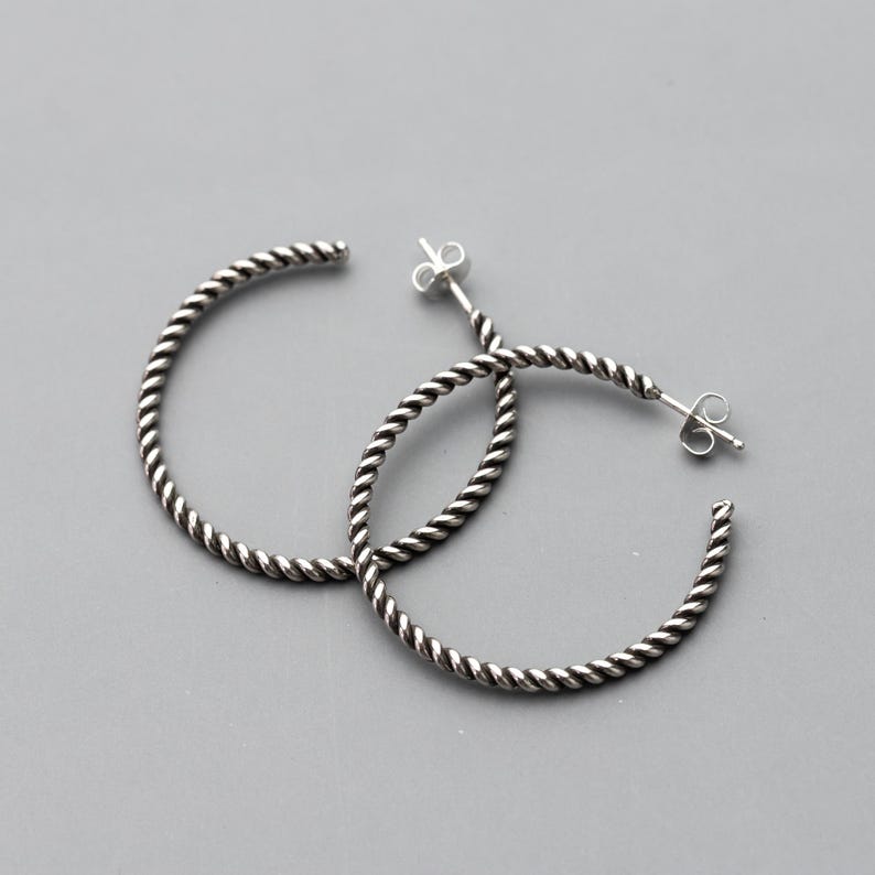 May include: A pair of silver hoop earrings with a twisted rope design. The earrings have a post and butterfly clasp closure. The hoops are a medium size and are displayed on a gray background.