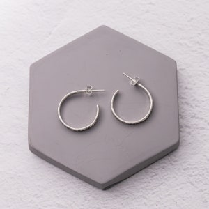 Pode incluir: Um par de brincos de argola de prata com acabamento texturizado. Os brincos s&atilde;o exibidos em uma superf&iacute;cie hexagonal cinza.