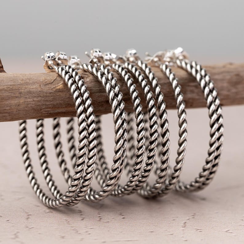 May include: A collection of silver hoop earrings with a twisted rope design. The earrings are displayed on a wooden branch against a neutral background. Each earring has a small, decorative stud at the top.