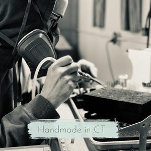 May include: A person wearing a respirator and glasses, soldering metal. The image is in black and white, with the text "Handmade in CT" in a light blue banner at the bottom.
