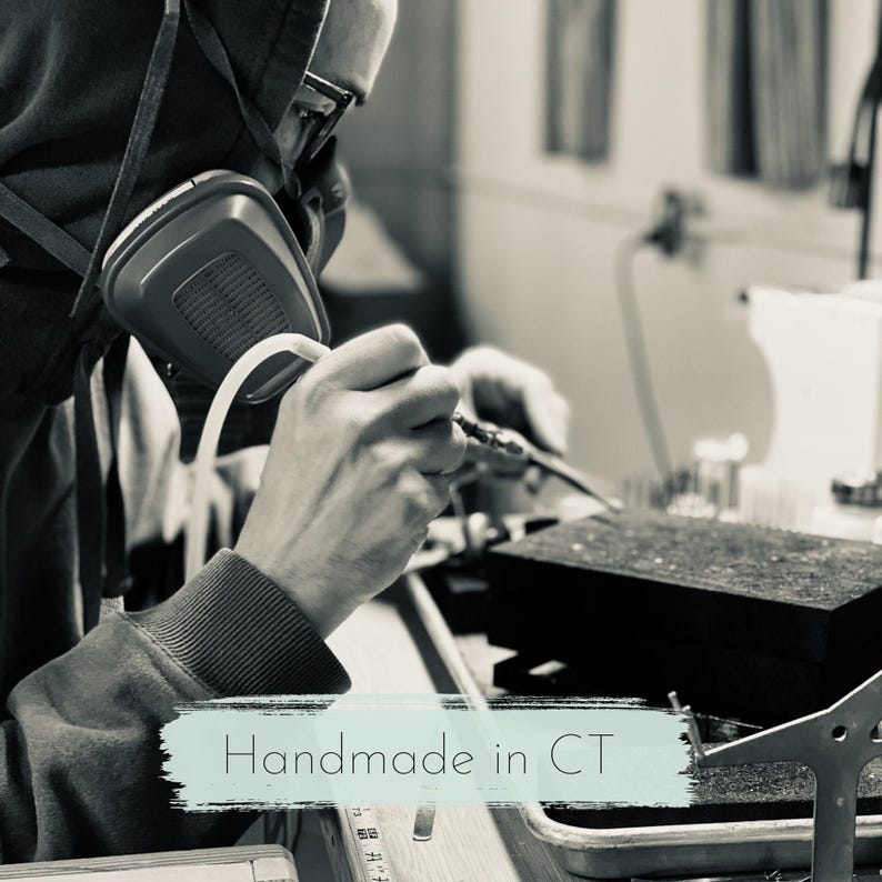 May include: A person wearing a respirator and glasses, soldering metal. The image is in black and white, with the text "Handmade in CT" in a light blue banner at the bottom.