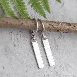 May include: A pair of silver rectangular earrings with a textured surface. The earrings hang from a silver hook and are displayed on a wooden branch against a gray background. The earrings have a hammered texture.